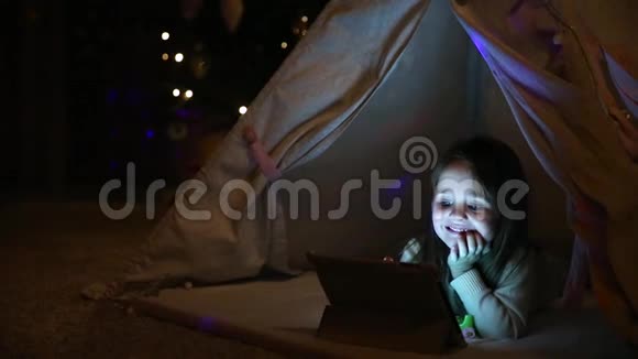 一个小女孩躺在地上看动画片坐在圣诞树背景下的帐篷里视频的预览图