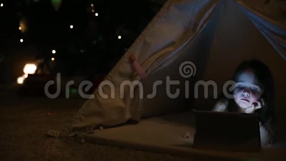 坐在家里的帐篷里一个躺在地上的小女孩在一棵圣诞树的背景下点击视频的预览图