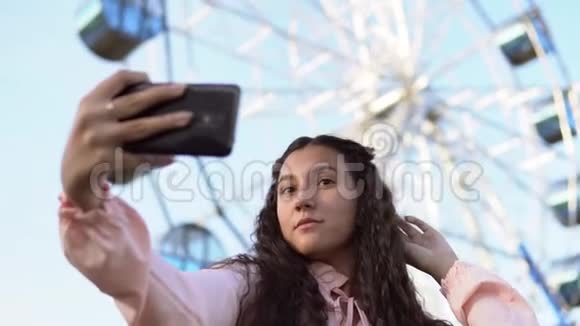 一个穿着连衣裙留着长发的女孩用手机在摩天轮附近自拍慢动作肖像视频的预览图