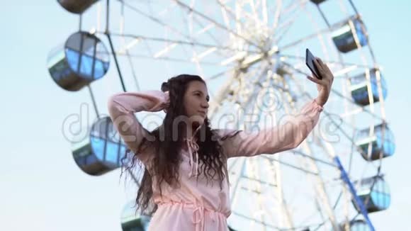 一个年轻的女孩留着长发穿着一件长裙用一部智能手机站在摩天轮附近自拍慢动作视频的预览图