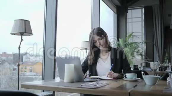 漂亮的女人在笔记本上做笔记年轻的女商人坐在木桌旁的咖啡店里桌子上是灰色的视频的预览图