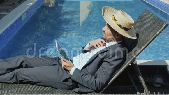 戴草帽的英俊商人在游泳池附近使用平板饮料和鸡尾酒视频的预览图