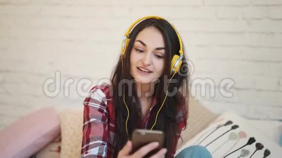 年轻女子带着耳机在智能手机上发短信那个女孩通过视频在电话里交谈穿黄色衣服的女人视频的预览图