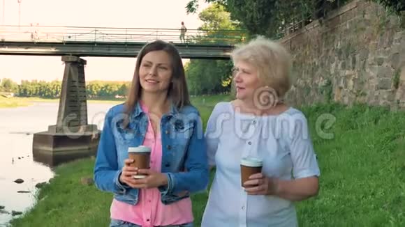 年长的女人和漂亮的年轻女孩沿着海滨散步喝咖啡野石的桥和老墙视频的预览图