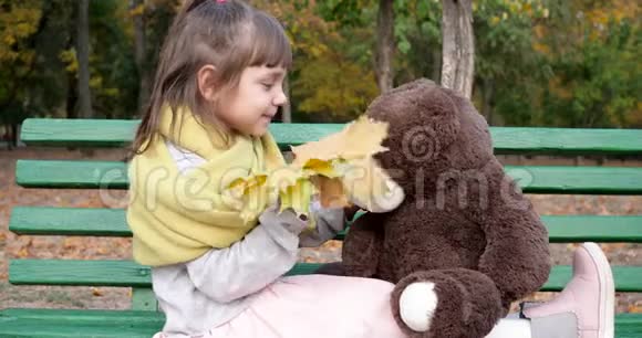 在公园的操场上一个拿着黄叶的小男孩在长凳上玩玩具熊玩具视频的预览图