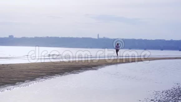 一个夏天的早晨穿着运动制服的年轻苗条的黑发女人沿着沙质海岸奔跑视频的预览图