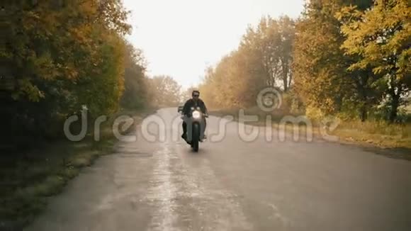 阳光明媚的森林里一个穿着太阳镜和皮夹克的时髦年轻人骑着摩托车在柏油路上视频的预览图