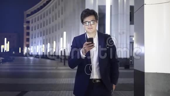 年轻的商人带着智能手机和耳机走在城市的一条夜晚街道上视频的预览图