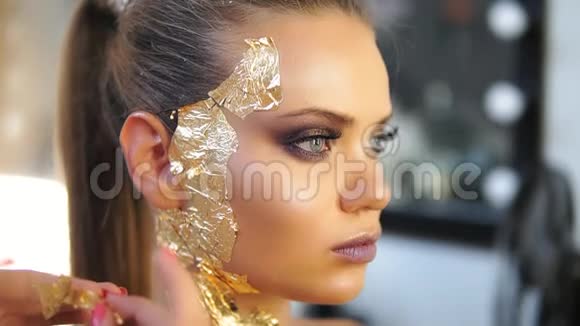 一位化妆师将金色闪亮的金属纸贴在模特脸上特写镜头为筹备视频的预览图