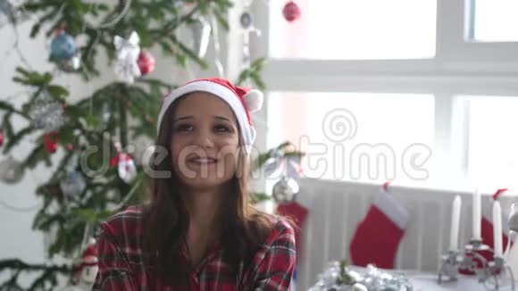戴着圣诞帽的快乐年轻漂亮的女人把纸屑扔在窗边的圣诞树旁并展示了它的形状视频的预览图