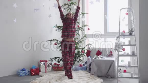 穿着睡衣的年轻黑发女人站在圣诞树附近把纸屑扔到窗边慢动作视频的预览图