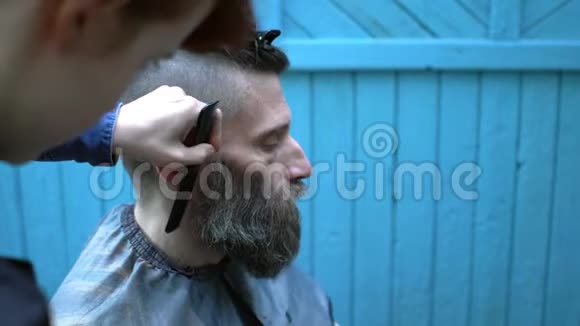 年轻女子理发师修剪奇怪的中年嘻哈男子在实验户外理发店的胡须男人看着视频的预览图