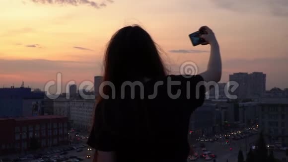 女孩在更漂亮的建筑上自拍从高楼的屋顶眺望市中心的傍晚视频的预览图