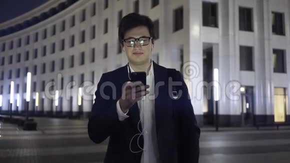 年轻的商人带着智能手机和耳机在一个夜城散步视频的预览图