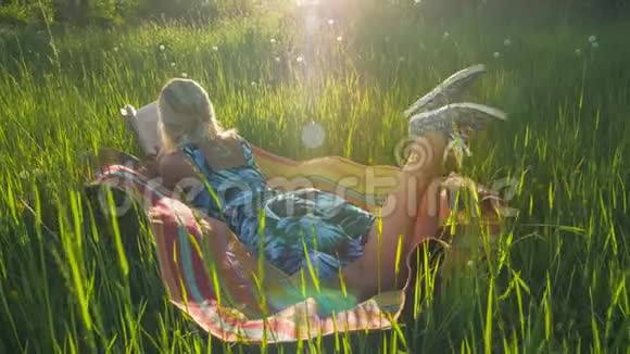 一个年轻的金发女孩躺在草地上的毯子上看书背光日落照明弹在框架后景手持视频的预览图
