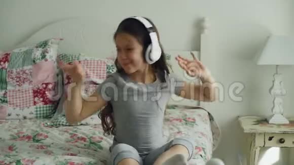 戴着耳机微笑着可爱的女孩坐在床上在家里跳舞很有趣视频的预览图