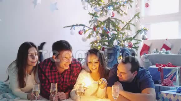 快乐的年轻朋友躺在圣诞树旁的地毯上在装饰好的房间里点燃火花庆祝新年视频的预览图