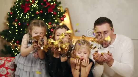 年轻幸福的四口之家坐在圣诞树旁吹着金色的纸屑可爱的母亲父亲和两个视频的预览图