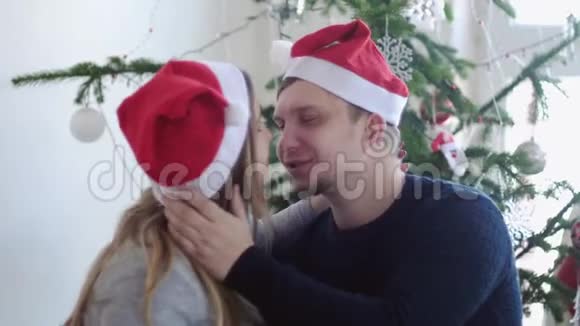 戴着圣诞帽的幸福年轻夫妇正在美丽的圣诞树旁接吻慢动作关门视频的预览图