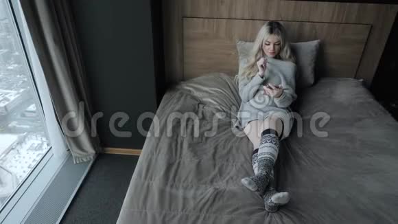 美丽的金发女人坐在床上带着智能手机女孩微笑心情好博客浏览互联网视频的预览图