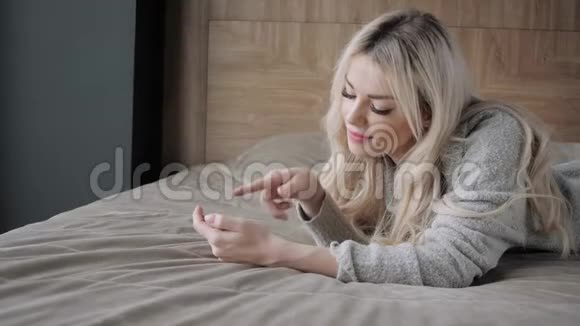 美丽的金发女人躺在床上带着智能手机女孩微笑心情好博客浏览互联网视频的预览图