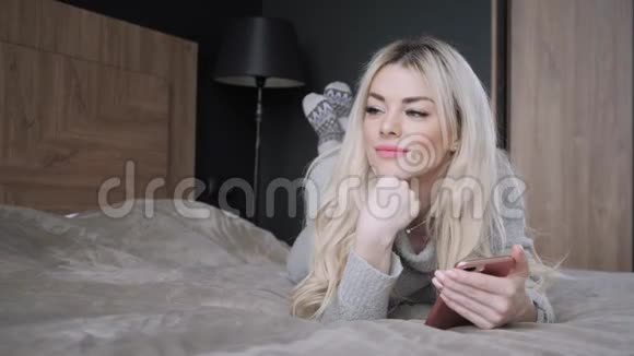 美丽的金发女人躺在床上带着智能手机女孩微笑心情好博客浏览互联网视频的预览图