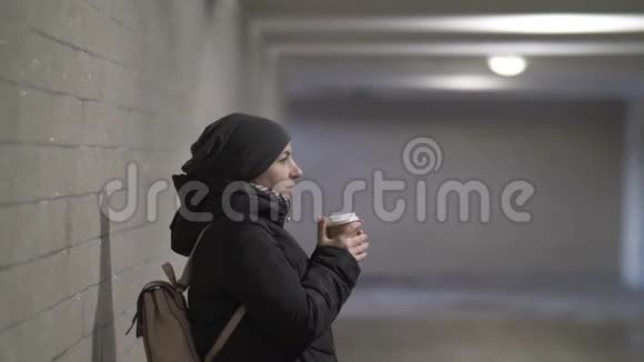 美丽的女人站在地下孤独的通道里享受着咖啡等待着某人冬天视频的预览图