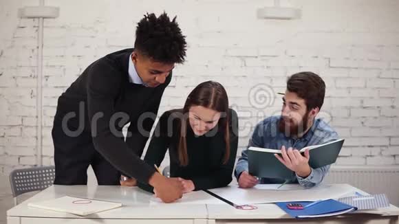 年轻女子正在用铅笔坐在桌子旁画一个计划她的非洲同事帮助她完成她的工作视频的预览图