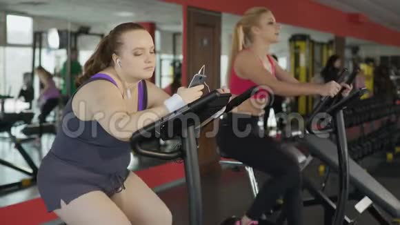 懒洋洋的超重女人在运动自行车上缓慢地骑着在电话上滚动视频的预览图
