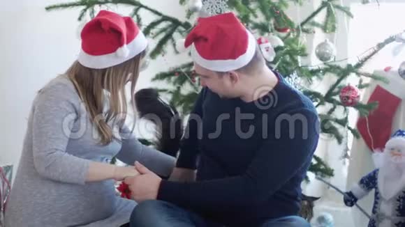 圣诞老人帽里快乐的年轻家庭丈夫抱着她怀孕的妻子靠近漂亮的圣诞树和大姑娘视频的预览图
