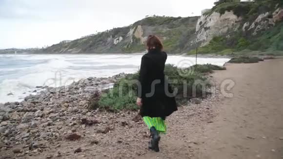 女人在海岸上散步穿着黑色衣服的女性走在海边多云风天气视频的预览图