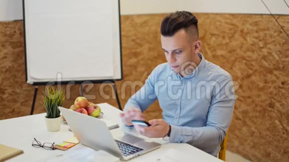 年轻的帅哥正在使用智能手机在舒适的办公室里的笔记本电脑上工作视频的预览图