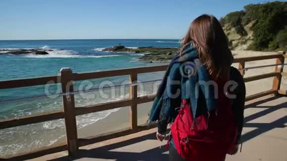 带背包和围巾的年轻女子的后景越来越接近海边的栅栏视频的预览图