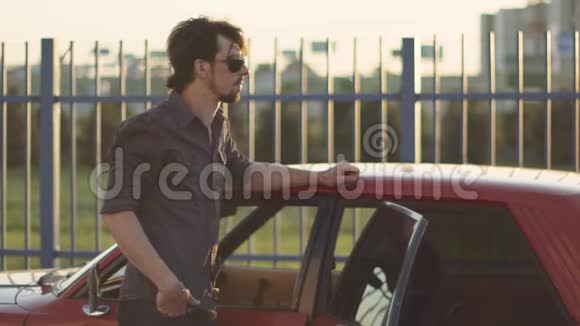 在日落或日出时英俊的男子和他那辆老式的强力汽车在街上的肖像视频的预览图