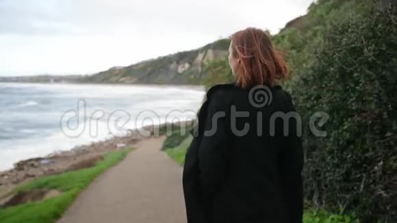 女人在海岸上散步穿着黑色衣服的女性走在海边多云风天气视频的预览图
