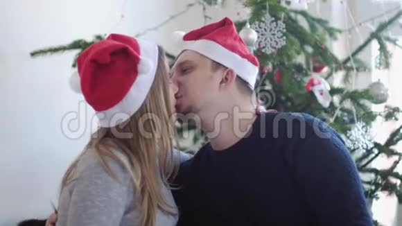 圣诞老人帽里快乐的年轻家庭丈夫亲吻他怀孕的妻子靠近美丽的装饰圣诞树slwo运动视频的预览图