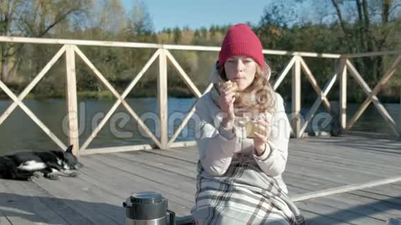 一位穿着保暖衣服的女游客在河岸附近的一座桥上背着背包喝着热茶野餐一只狗躺着视频的预览图
