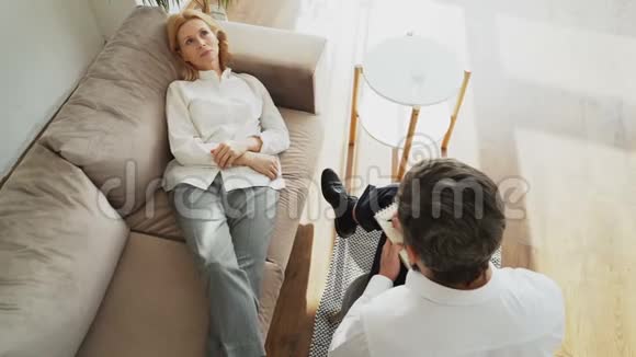 躺在沙发上与专业男性心理治疗师或心理学家谈论她的问题的沮丧女人的最高看法视频的预览图