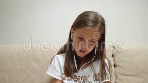 一个8岁的女孩坐在沙发上用智能手机用耳机听音乐视频的预览图