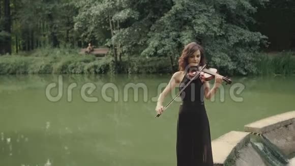 穿着黑色连衣裙拉小提琴呆在桥上户外的年轻漂亮女孩视频的预览图