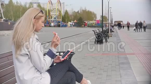 一位年轻漂亮的金发女郎在河堤的长凳上放松时吃了一个甜甜圈享受着她的智能手机视频的预览图