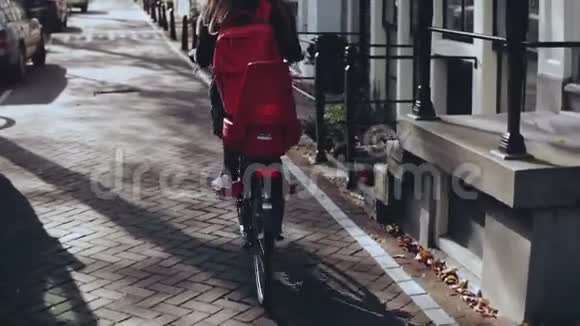 旅游女孩骑自行车在阳光明媚的城市街道后视慢动作骑着红色背包的女人骑着自行车巡航视频的预览图