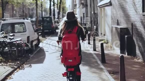 后景女性骑着阳光明媚的城市自行车慢动作穿着时髦帽子的女孩骑自行车背着红色背包探索视频的预览图