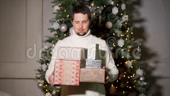一个穿着毛衣在圣诞树背景上放有礼物的盒子的快乐男人的肖像视频的预览图