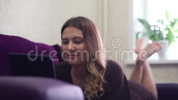 漂亮的年轻女孩用平板电脑躺在沙发上视频的预览图