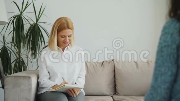 女性经验丰富的心理学家在办公室里谈论和倾听心烦意乱的女病人并在剪贴板上写笔记视频的预览图