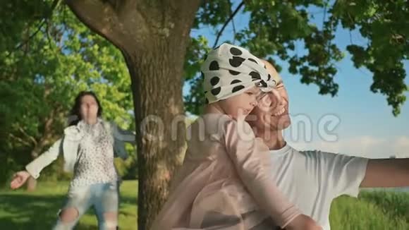 一个年轻的家庭和一个小女儿在湖边的一棵树下的公园里父亲把女儿抱在怀里视频的预览图