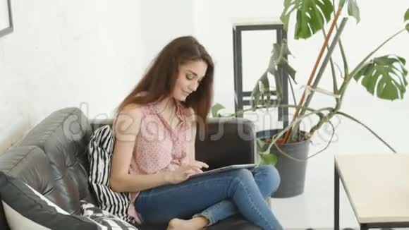 年轻的高加索女人坐在沙发上使用平板电脑视频的预览图