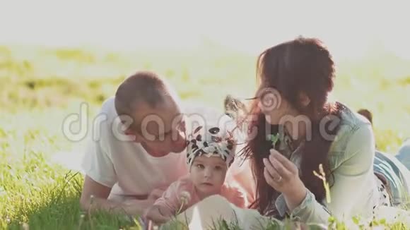 年轻快乐的父母和一个小可爱的女儿正躺在夏日阳光明媚的草地上吻视频的预览图