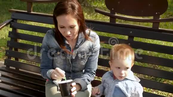 年轻的女人和孩子正在长凳上的公园里吃冰淇淋年轻的母亲用勺子喂儿子冰淇淋视频的预览图